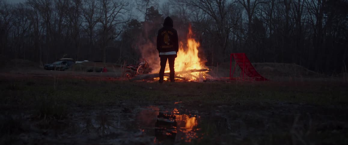Person wearing hoodie stands next to bonfire and red ramp occupying field between pond and leafless trees