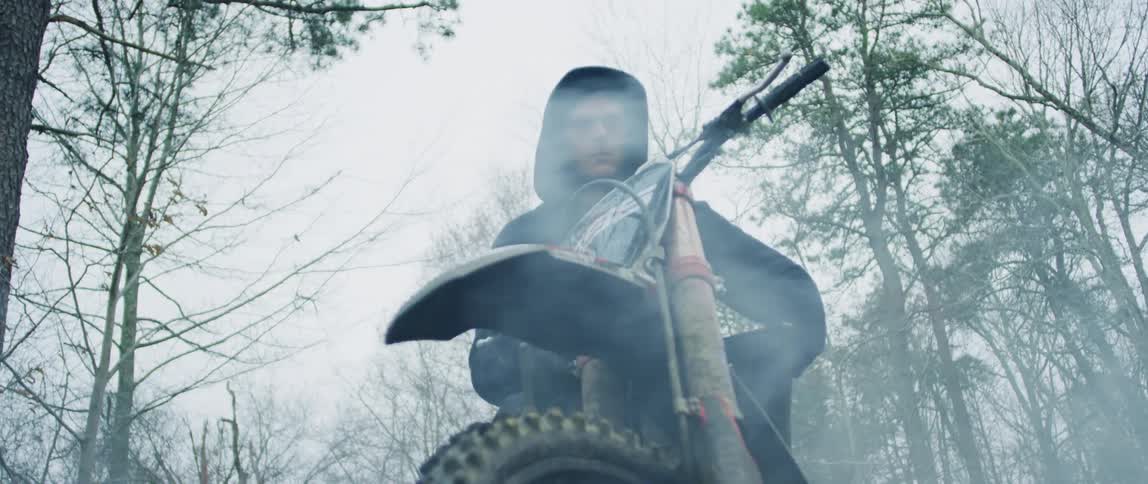 Man sits on motorcycle among conifers and leafless trees as smoke rises from the ground nearby