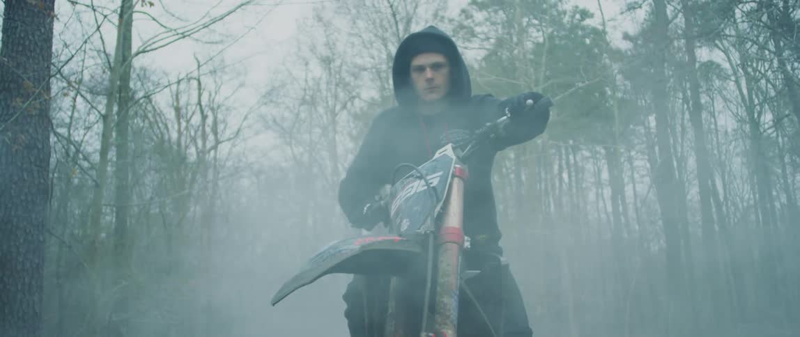 A man with a clenched jaw sits on a motorcycle in a forest as fog rolls over