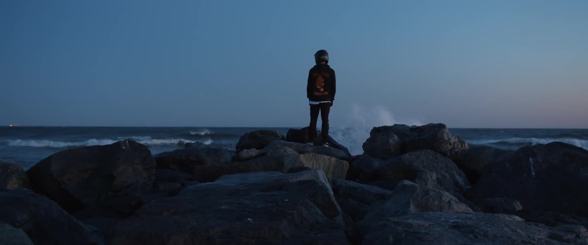 Person wearing crash helmet stands on boulders on shore looking out to sea
