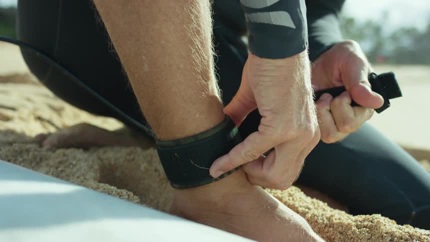 A surfer strapping a surfboard to their ankle on a sandy beach