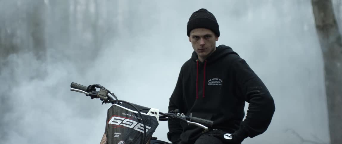 Young intense man standing over a parked motorcycle surrounded by white smoke in the forest