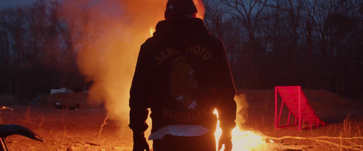 A man walks up to a fire burning outside and throws a liquid on the fire which causes the flames to raise even higher