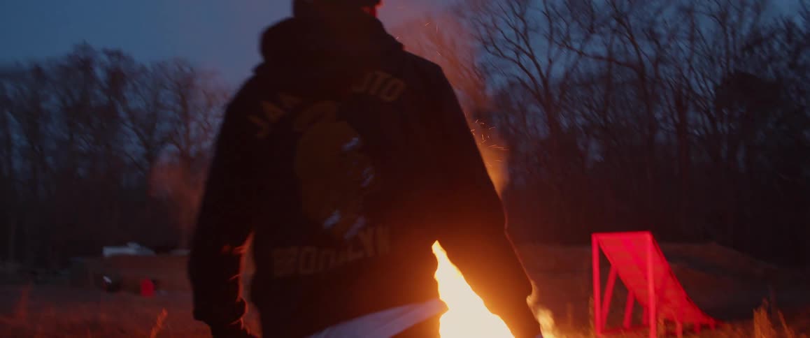 Man walks past motorcycle to add fuel to bonfire near ramp