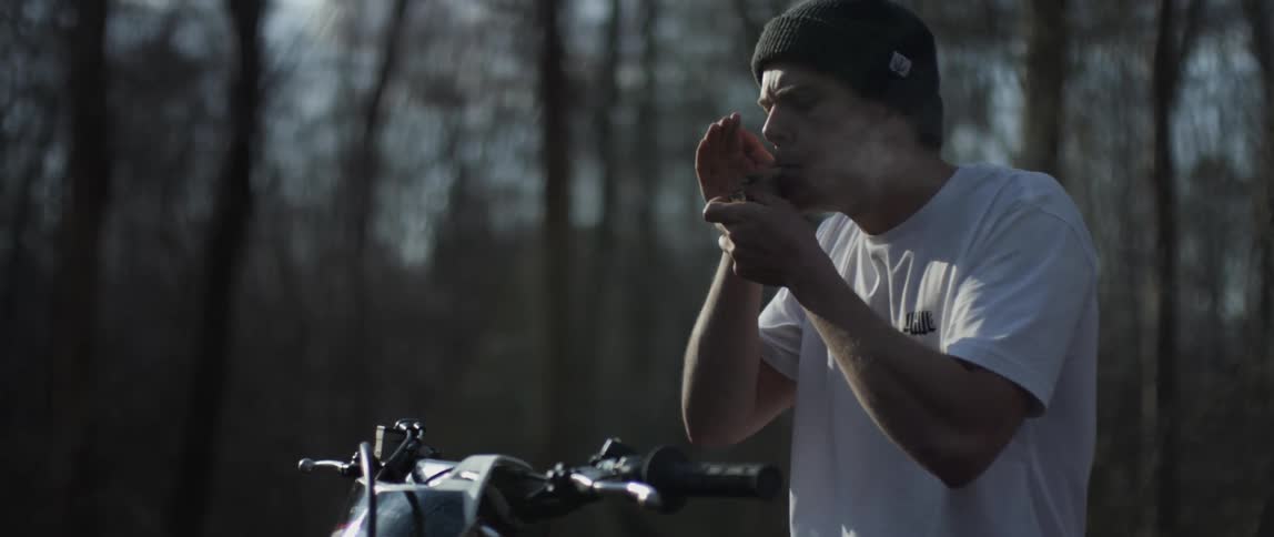 A man sitting atop a motorcycle in a woodland lights a cigarette