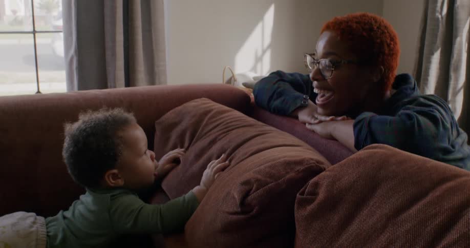 Mother looks over sofa back to son crawling on sofa and smiles
