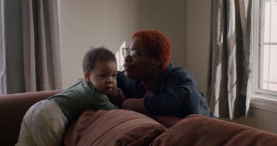 A woman kisses a toddler on the forehead as he climbs on a sofa