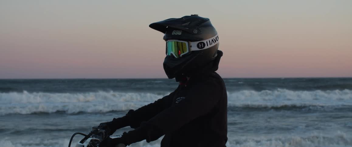 A person sitting on a motorcycle parked on a beach at sunset turns to look into the camera