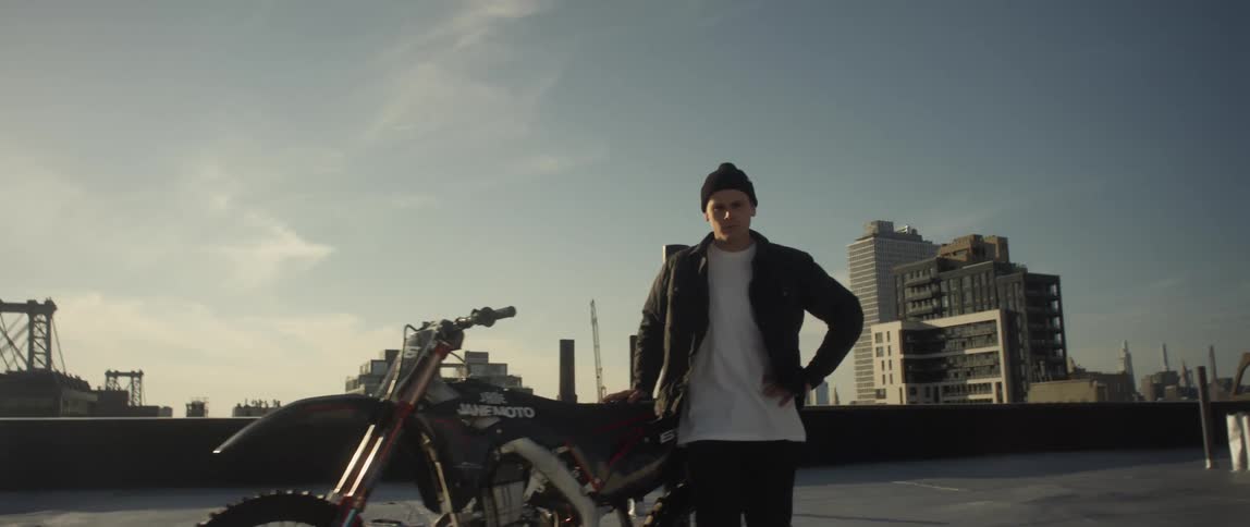 Glaring man stands next to motorcycle on rooftop with suspension bridge and city buildings in the background