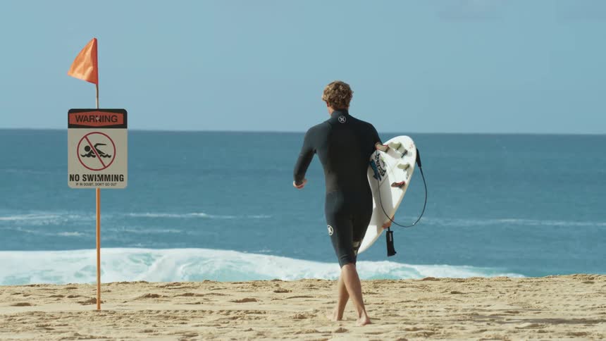 A surfer walks towards the ocean while carrying a surfboard before stopping near a sign prohibiting swimming