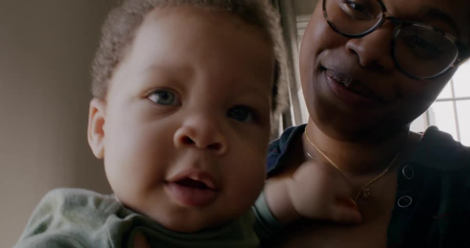 Mother holds baby and smiles as he grabs her necklace with one hand and reaches out with the other