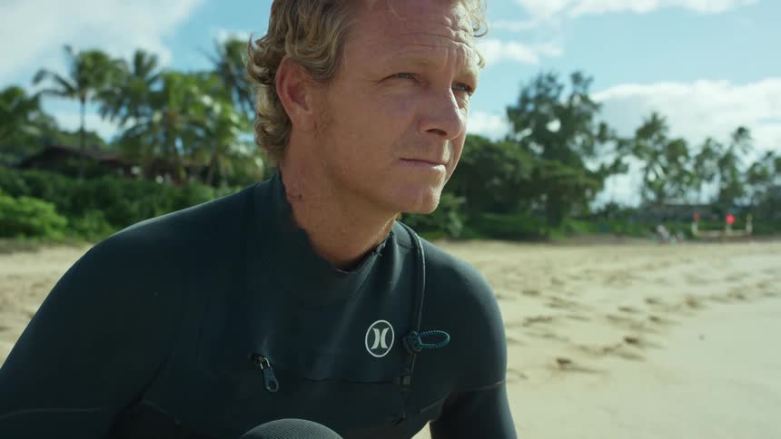 A male surfer is kneeling on a beach and strapping on a surfboard ankle leash
