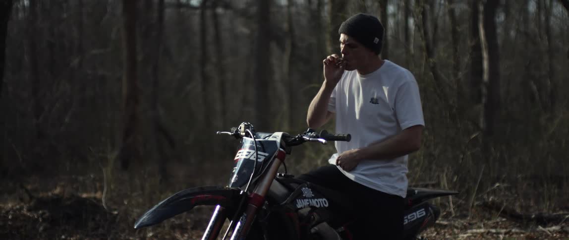 A man lights and smokes a cigarette while sitting on a motorcycle parked in a woodland during the day