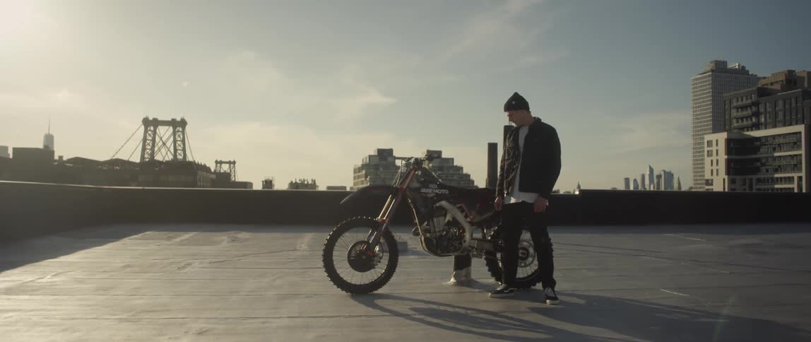 A man stands on an empty rooftop parking lot looking down at his bike