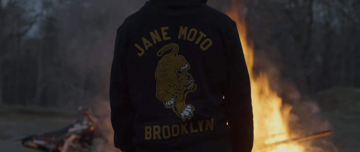Person with brand name jacket stands watching bonfire in field near leafless trees