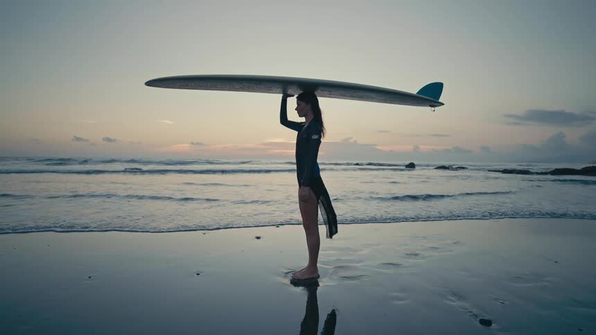 A woman is standing on the coast of a beach with a surfboard