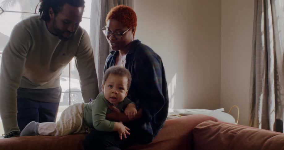 Mother holds baby as she sits on top of couch with father leaning close