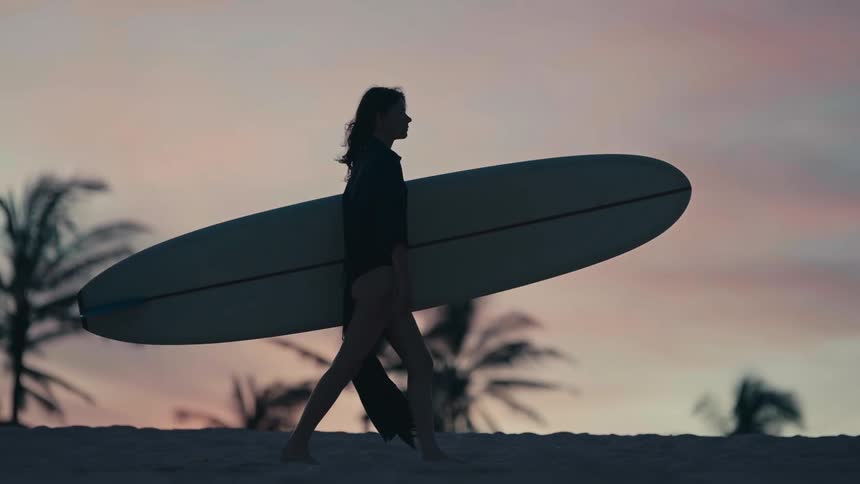 Silhouette of a woman carrying a longboard walking on the sand at dusk