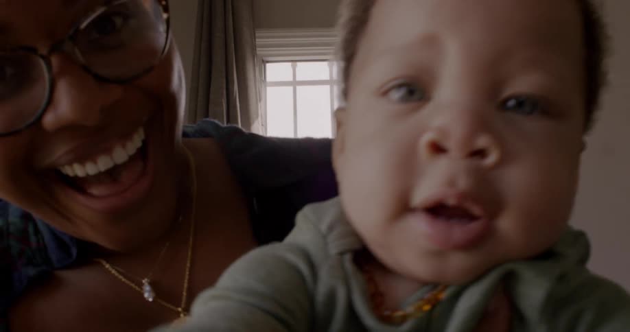 A mother smiles while holding her infant son as he reaches for items