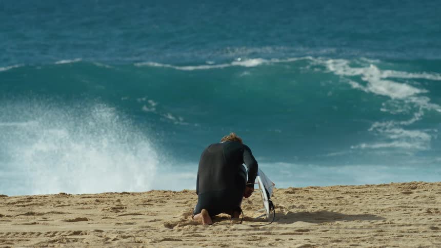 A man in a wet suit kneels down on the beach beside his surfboard