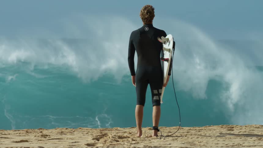 A man stands up beside an large incoming ocean wave holding a surfboard