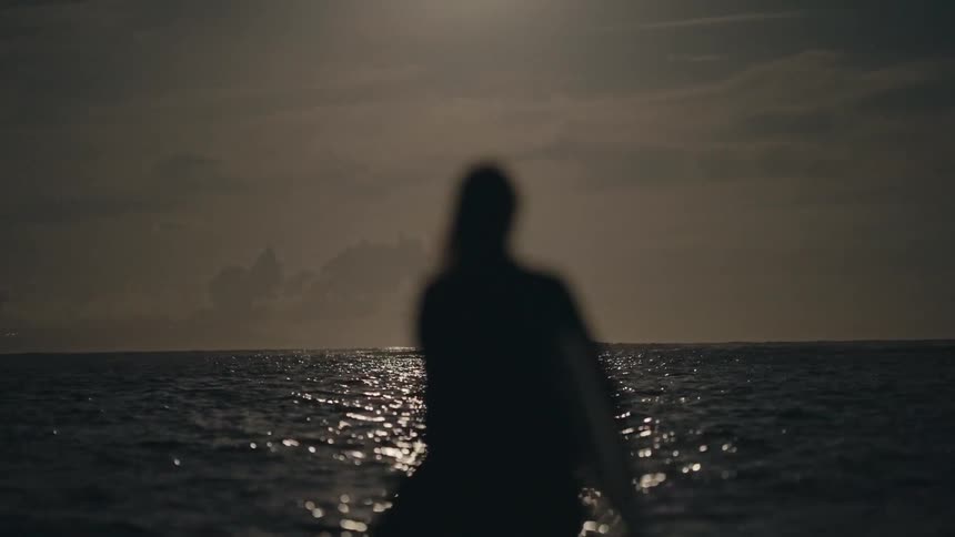 A surfer carries their surfboard into the water as it shines in the moonlight and there is a full moon in the sky