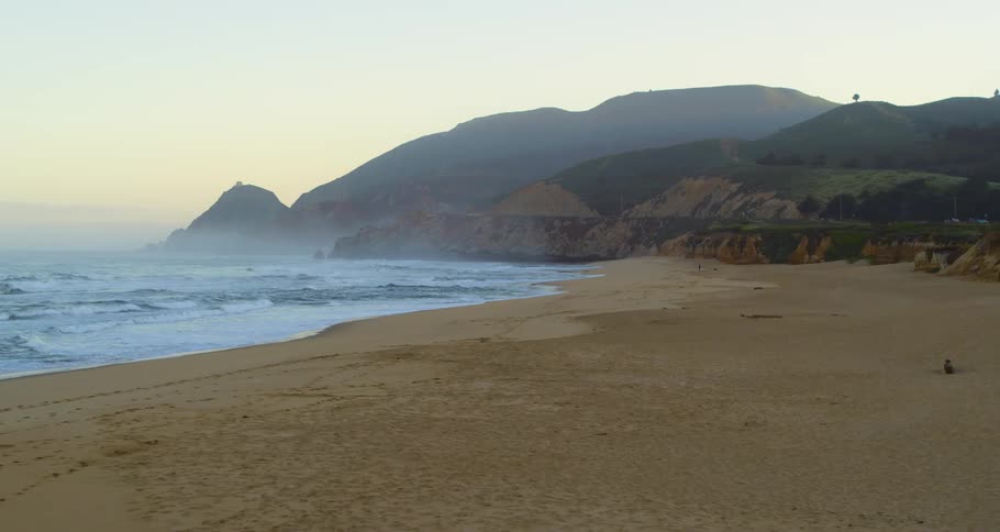 Waves roll onto beach sand as fog covers red tinted sky and promontory