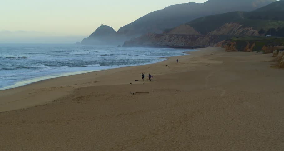 An aerial view of two surfers heading to the water on a beach while another man is walking with his dog during the day
