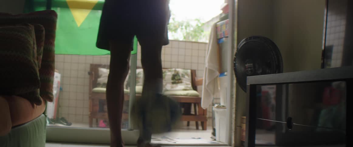 A young man juggles a soccer ball with his feet while standing in front of a brazilian flag indoors during the day
