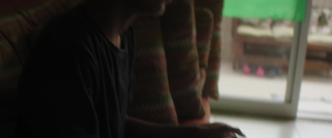 A young boy sits on a couch at home and plays a video game as someone kicks a ball in front of him