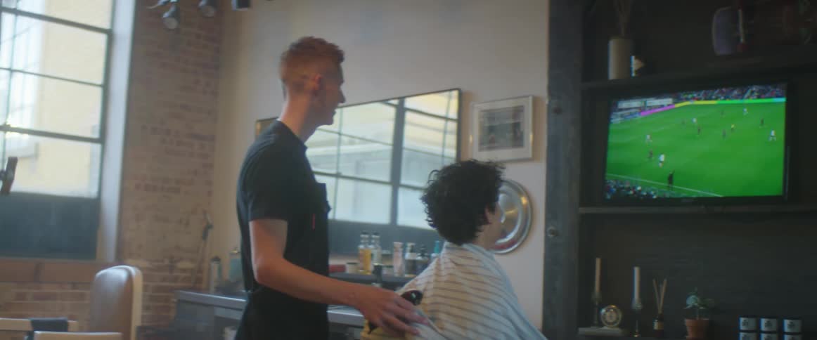 A barber holding a razor stands behind a clients chair as they both watch a soccer game on television
