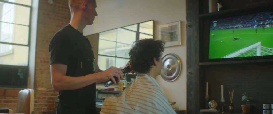 A man is trimming another mans hair as they watch a soccer game on the television