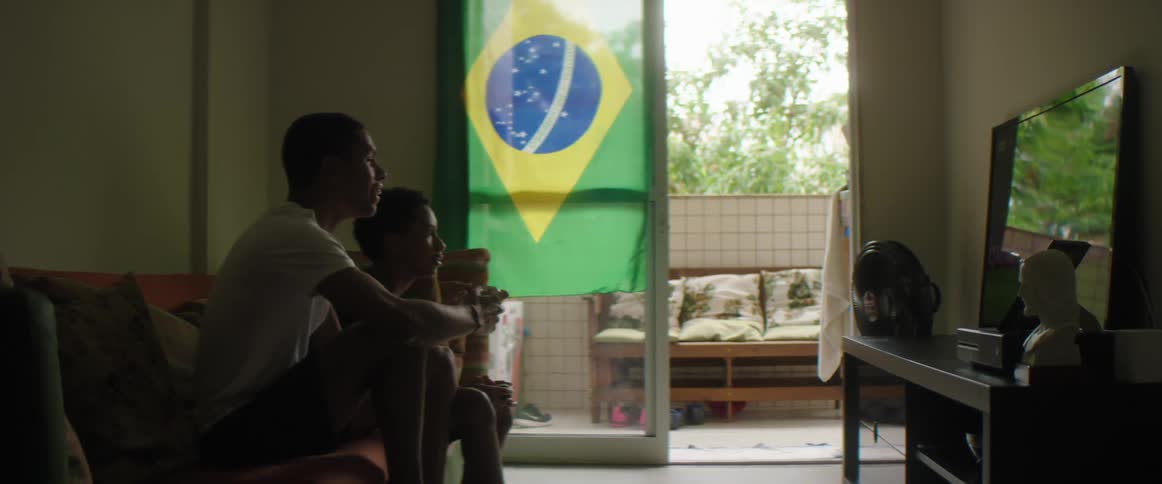 Two teen boys play video games sitting on couch next to brazilian flag hanging on the door