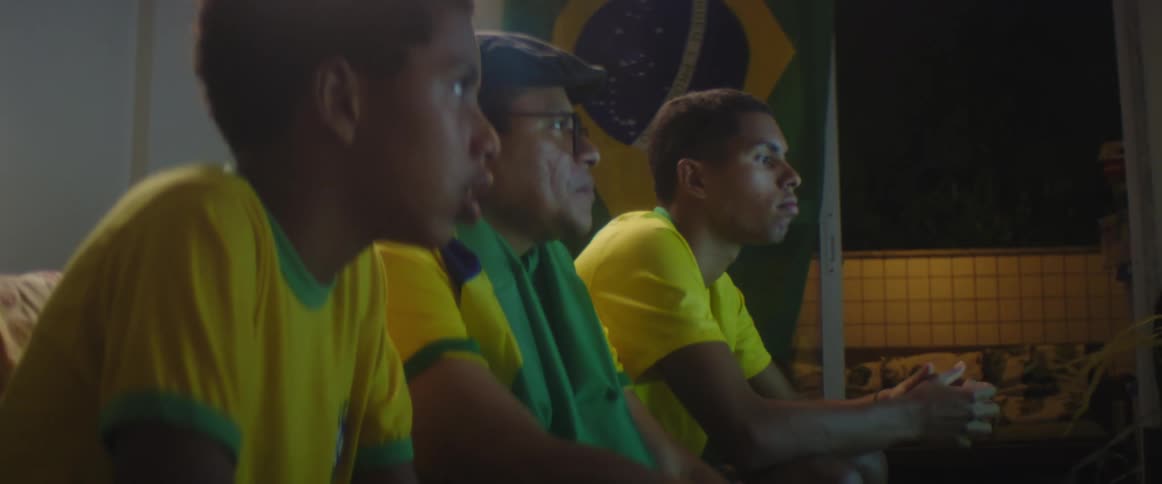 Father and sons wearing brazilian football uniform watch football match on television in living room at home