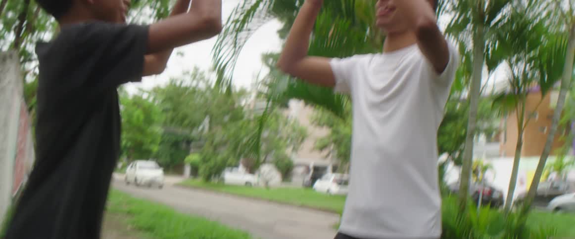 A pair of boys high five one another while standing over a trash bin used as a soccer goal on a neighborhood sidewalk during the day