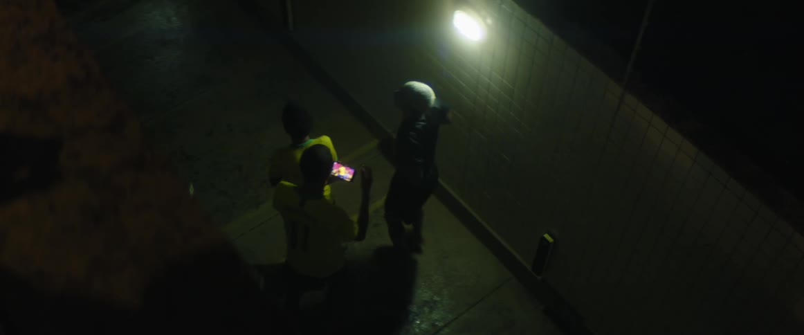 A group of young men cheer while watching sports together on a smartphone under a light in a dark alleyway at night