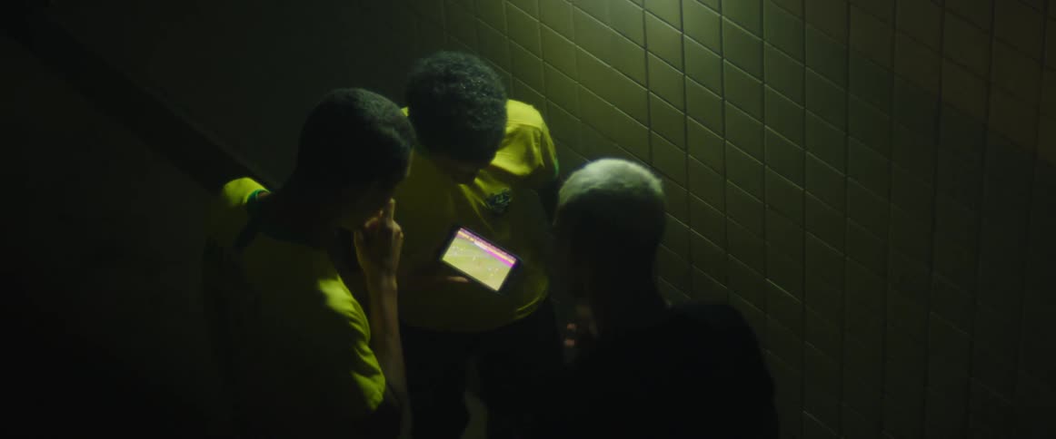 Teen boys wearing brazilian uniform watch football match on smartphone next to wall in the dark
