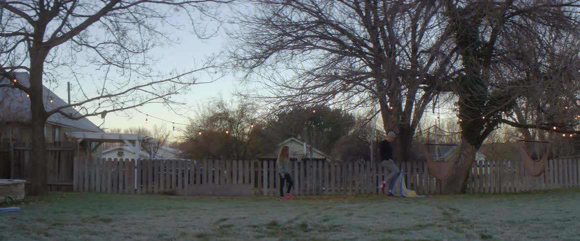 Father with gray beanie pants and black sweater plays football with blonde daughter with red shoes black pants and green shirt in the backyard
