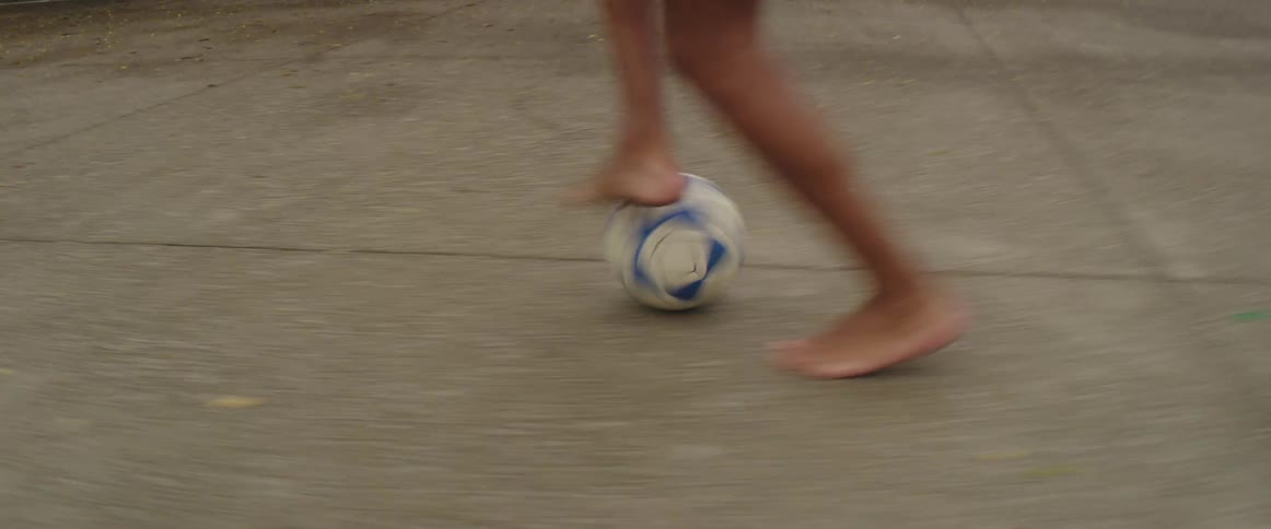 A barefoot soccer player kicking a ball into a goal on an outdoor court