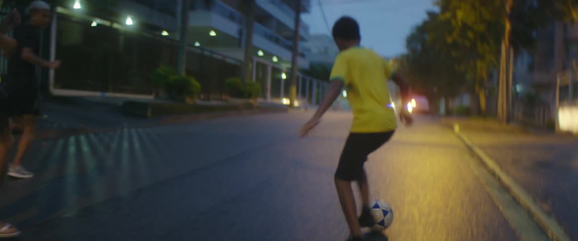 Three men kick a soccer ball down a neighborhood street as cars pass by