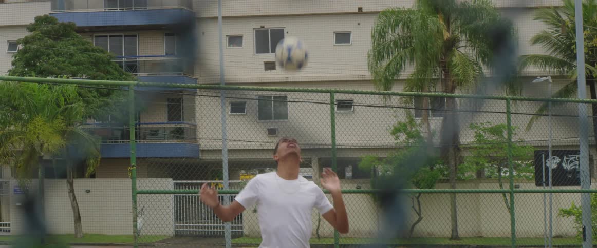 A teenager bounces a soccer ball on his head in a fenced area of a neighborhood