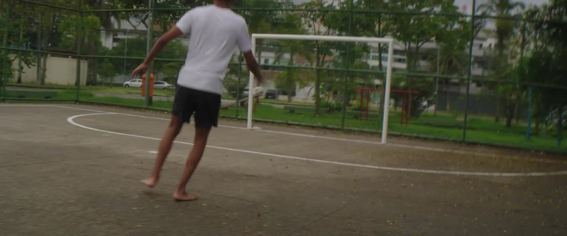 A boy is running barefoot on a soccer field kicking a ball into the goal