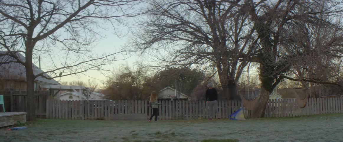 Father with gray beanie pants and black sweater plays football with blonde daughter with red shoes black pants and green shirt in the backyard