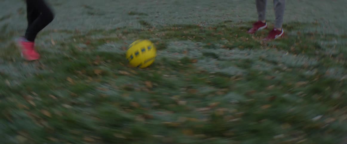 Father with gray beanie pants and black sweater plays football with blonde daughter with pink shoes black pants and green shirt in the backyard