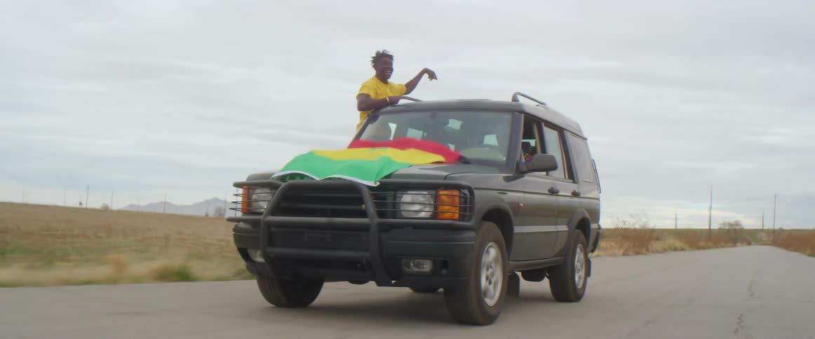 Young man celebrates on the window of a car driving on a road with a waving flag on the hood