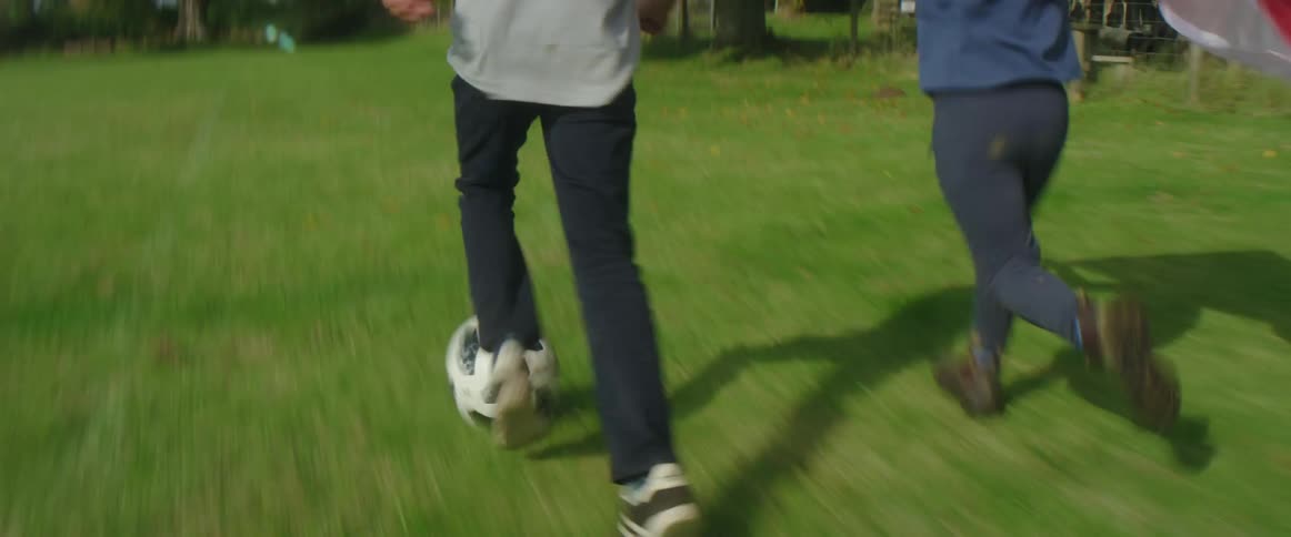 A young boy with a flag held behind his head runs across a field next to a boy kicking a soccer ball