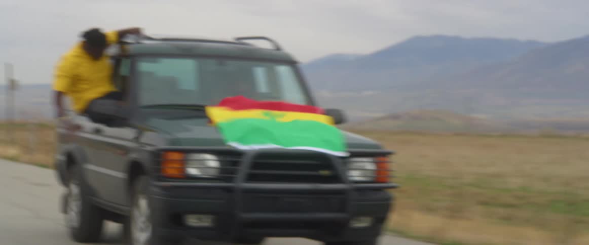 A man hangs out the window of a vehicle with a flag on the font and yells and cheers as another man drives