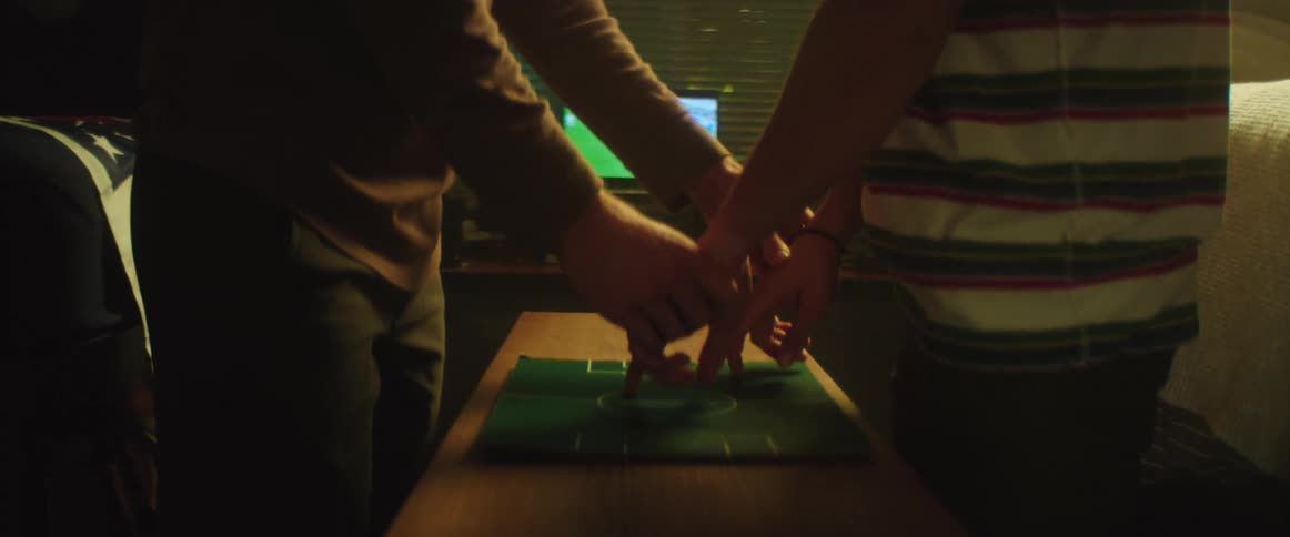 Friends holding hands while playing over a tabletop game celebrate and hug in front of a screen showing a soccer game