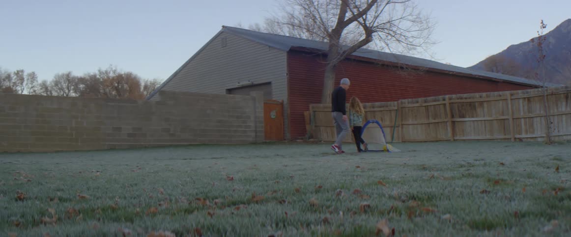 Father with gray beanie pants and black sweater plays football with blonde daughter with pink shoes black pants and green shirt in the backyard