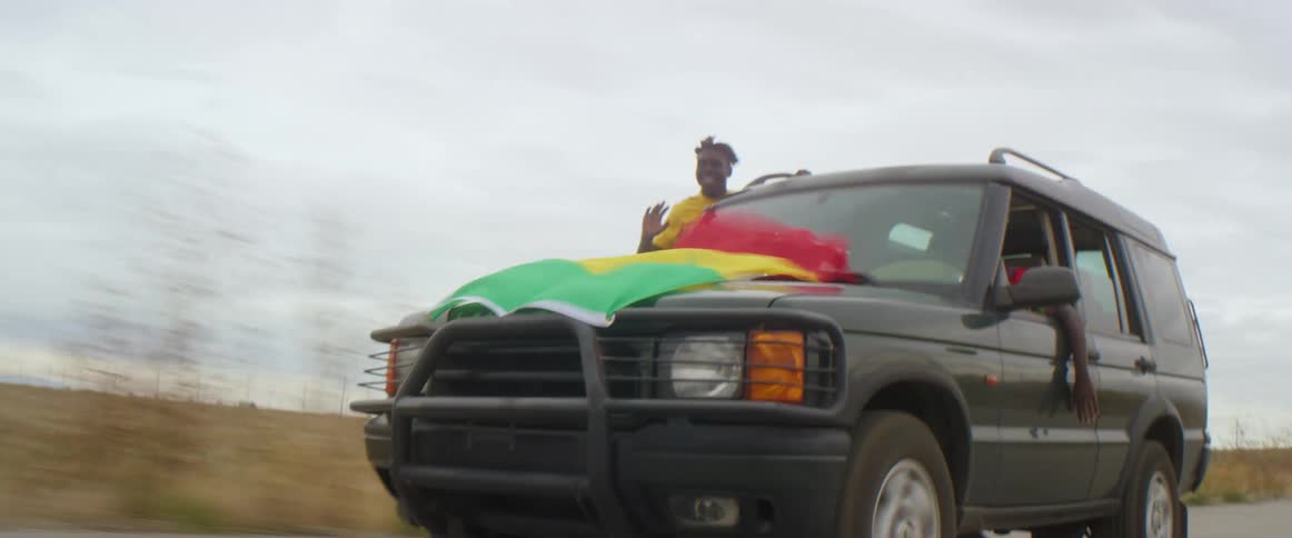 A man is speeding down a highway in an suv while another man is sitting on the passenger side door hanging out